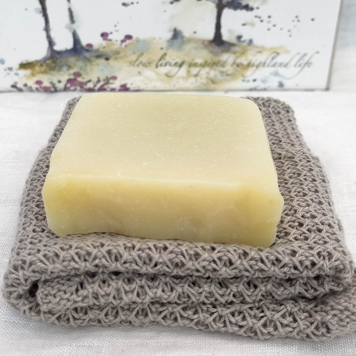 a solid soap bar sat on a handmade linen face cloth. the solid soap bar is called fresh facial bar and is handmade in Scotland by Made in the Glen. It is a natural soap and a lovely creamy colour. The linen face cloth is an oatmeal colour