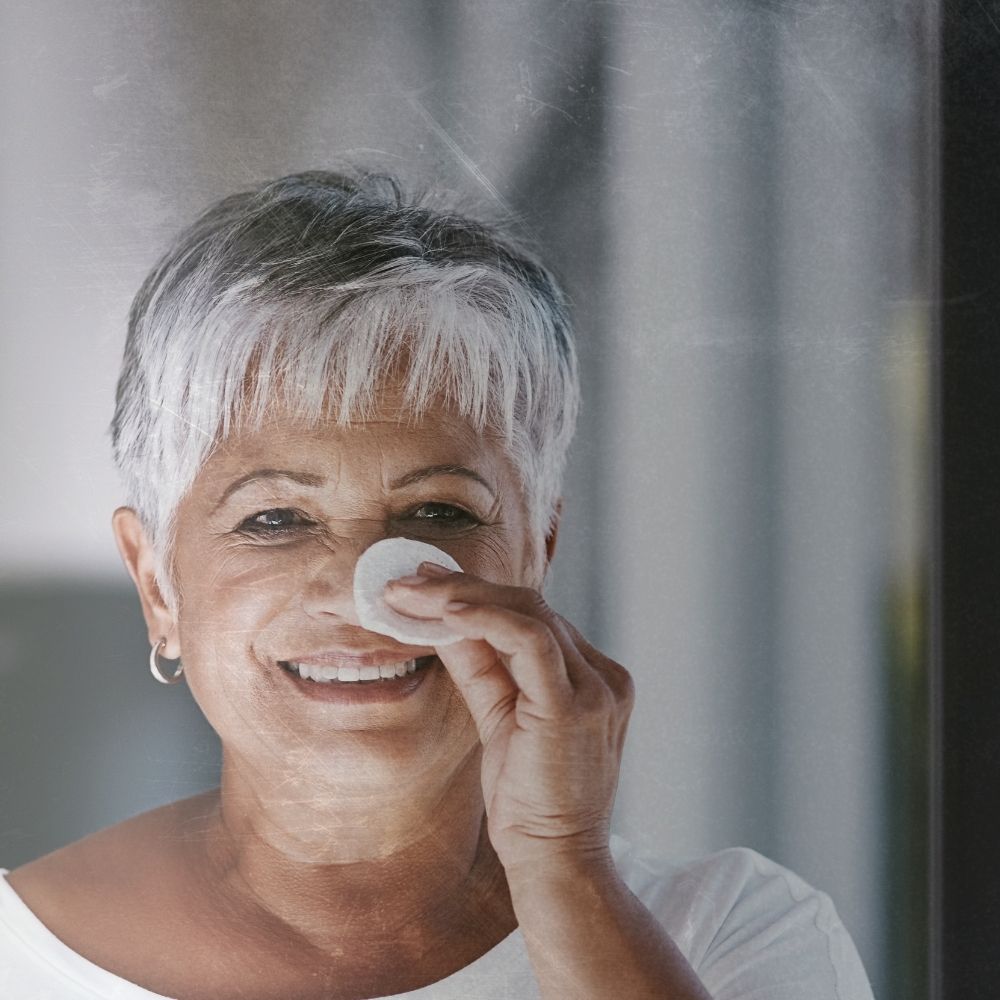 an older lady with greying hair is stood in front of the mirror she has a cotton pad in her hand and is wiping toner across her face she is using apple cider vinegar with the mother in it as a facial toner