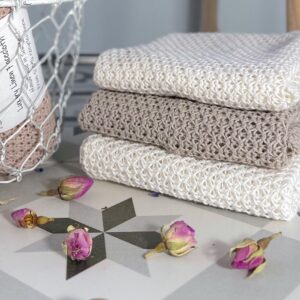 a stack of linen face cloths that have been handmade in Scotland. Two white and an oatmeal in front of the stack of face cloths are a few dried rose buds. off to the side and slightly out of sight is a metal wire basket with more face cloths in it.