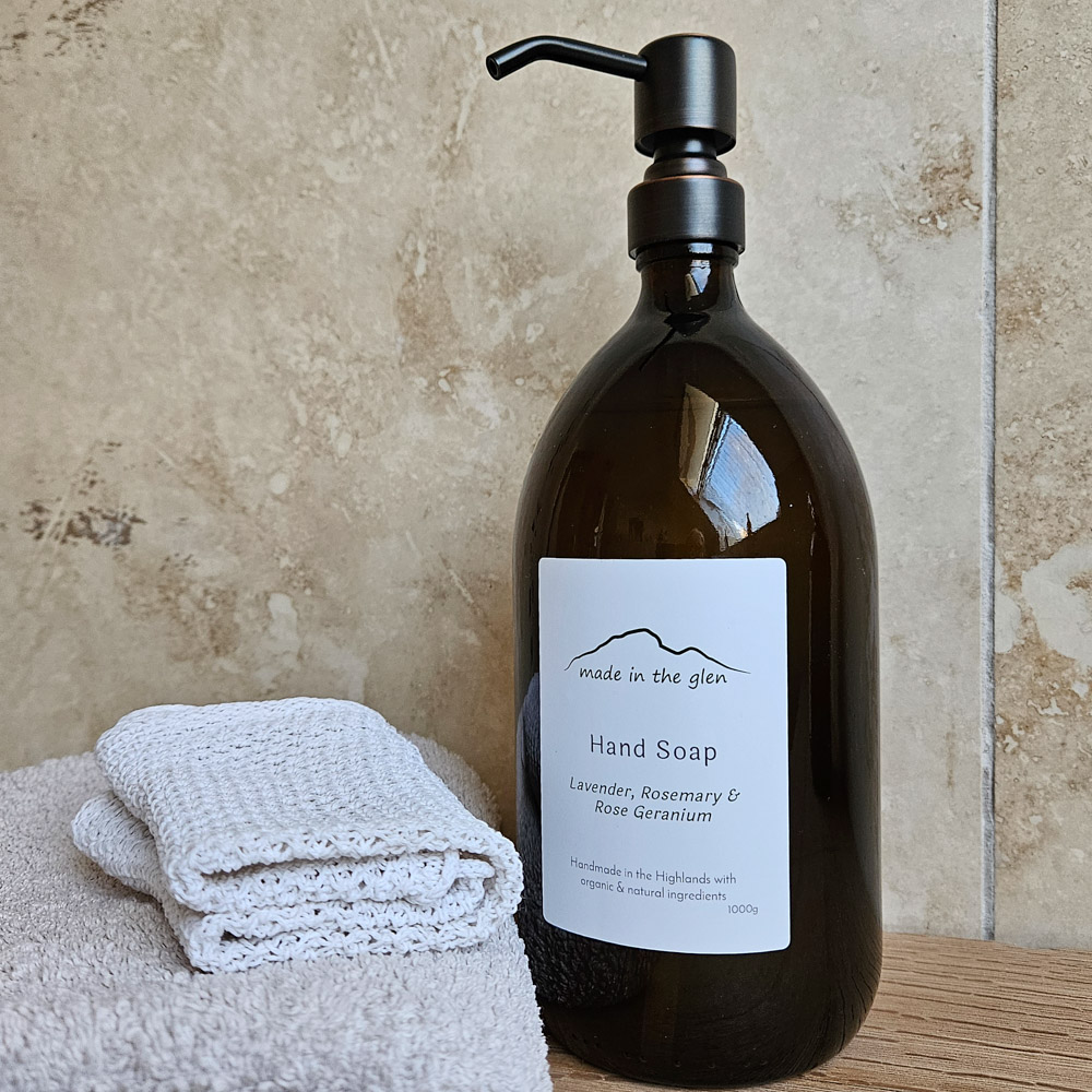 Amber glass bottle with brass metal pump filled with Made in the Glen Liquid hand soap made with organic ingredients on an oak worktop in the batheroom bed=side it is a bar of handmade soap and two white bottles of Ola Ròs Skincare products