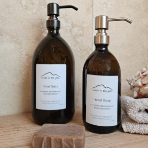 Two liquid hand soaps by Made in the Glen Lavender, Rosemary 7 Rose Geranium Organic essential oils in an Amber glass bottle with brass metal pump filled with Made in the Glen Liquid hand soap made with organic ingredients on an oak worktop in the bathroom beside it is a bar or handmade soap