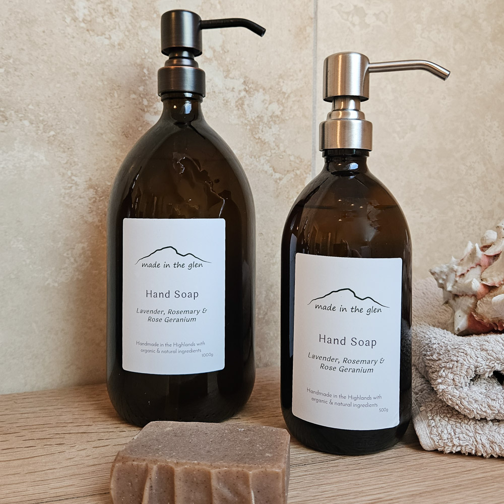 Two liquid hand soaps by Made in the Glen Lavender, Rosemary 7 Rose Geranium Organic essential oils in an Amber glass bottle with brass metal pump filled with Made in the Glen Liquid hand soap made with organic ingredients on an oak worktop in the bathroom beside it is a bar or handmade soap