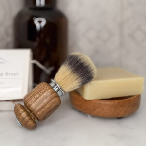 a traditional shaving brush laid on its side, with its brush resting on a bar of soap, the soap is on a handmade soap dish. the shaving brush was handmade in Scotland by Made in the Glen. the background is tiled and a dark amber regents style bottle stands behind the shaving brush and soap.