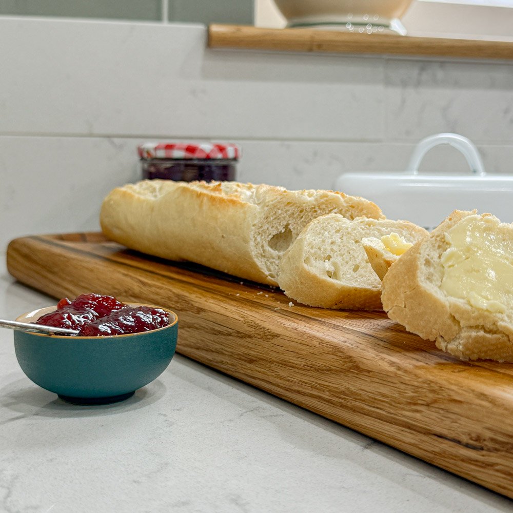 whisky cask grazing or garlic bread board for tearing and sharing with a baguette on it with a pot of bonne maman jam behind and an enamel white butter dish set on white marble kitchen worktop