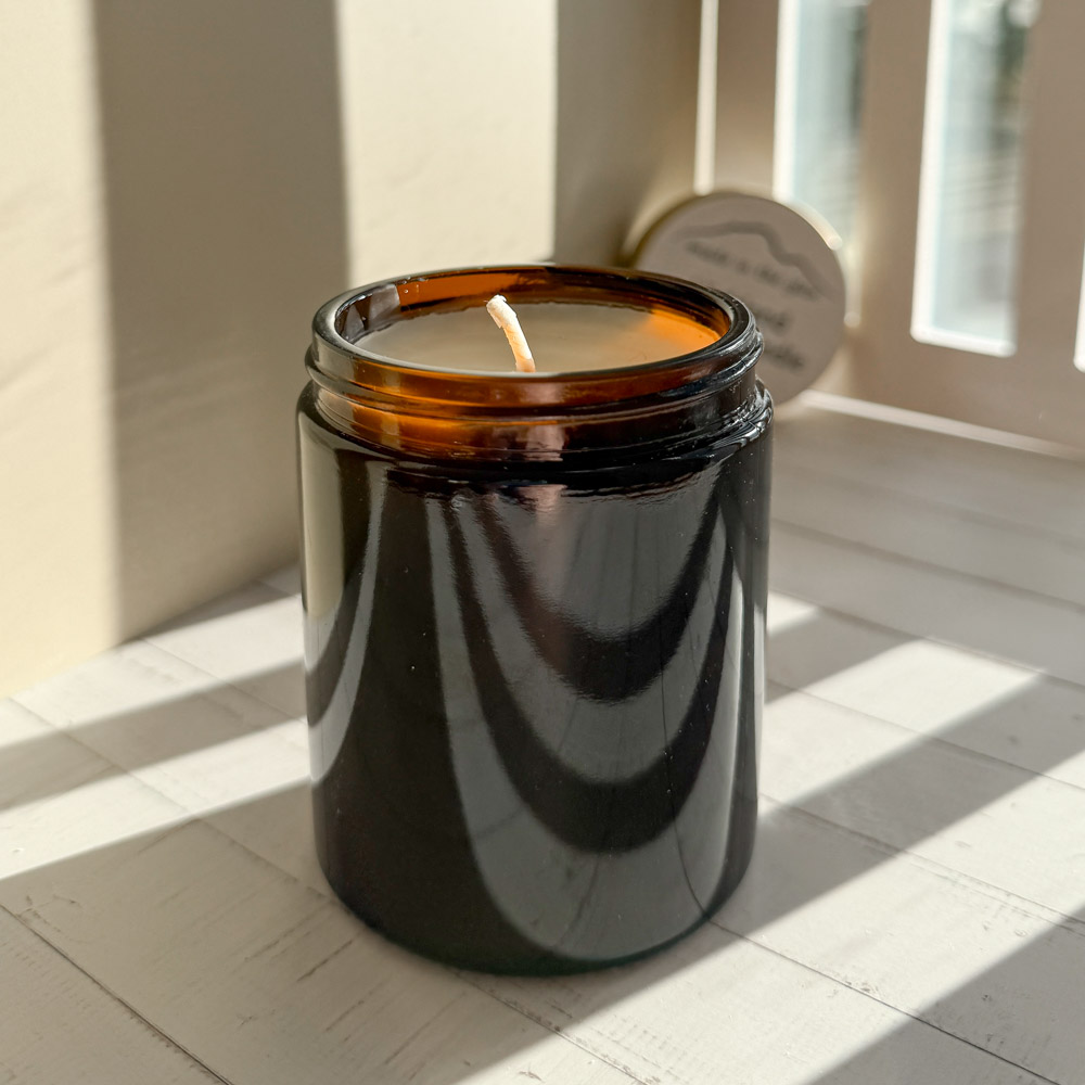 An amber glass jar scented candle with a piece of pine branch laid beside it. The scent of the eco soy wax candle with eco wick is highland forest, evoking memories of a walk in the forest in scotland