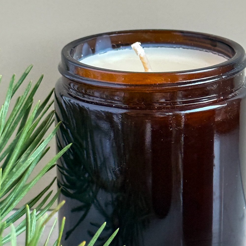 An amber glass jar scented candle with a piece of pine branch laid beside it. The scent of the eco soy wax candle with eco wick is highland forest, evoking memories of a walk in the forest in scotland
