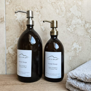 Two liquid hand soaps by Made in the Glen sweet orange & cinnamon Organic essential oils in an Amber glass bottle with brass metal pump filled with Made in the Glen Liquid hand soap made with organic ingredients on an oak worktop in the bathroom beside it is a bar or handmade soap