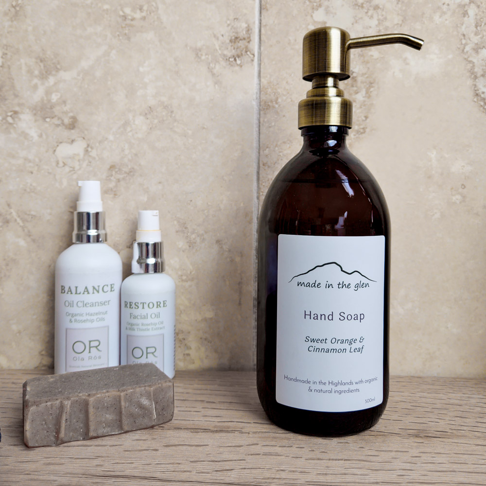 Amber glass bottle with brass metal pump filled with Made in the Glen Liquid hand soap made with organic ingredients on an oak worktop in the batheroom bed=side it is a bar of handmade soap and two white bottles of Ola Ròs Skincare products