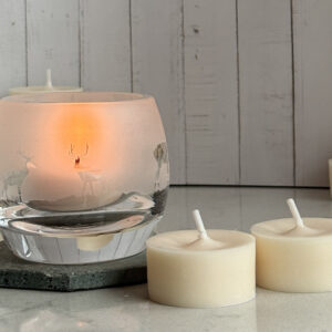two naked tea lights on a table top with a glass carved with a highland stag scene next to it the glass is a tea light holder and has a lit tea light inside it is giving off an orange glow. a wooden clad background is behind.