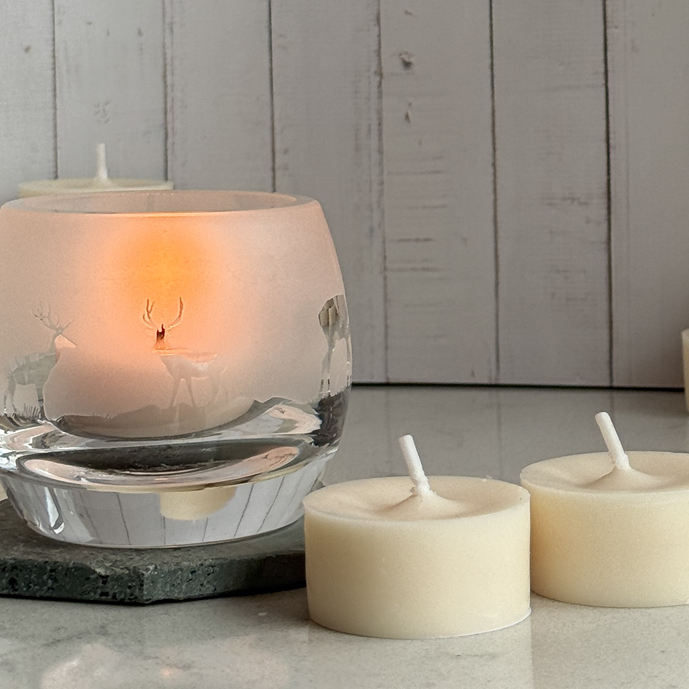two naked tea lights on a table top with a glass carved with a highland stag scene next to it the glass is a tea light holder and has a lit tea light inside it is giving off an orange glow. a wooden clad background is behind.