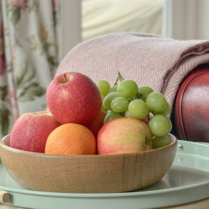 natural oak serving board filled with fresh fruit, apples, oranges and grapes, the bowl is on a small metal side table next to a leather chesterfield chair, a pink wool blanket is laid neatly over the arm of the chair, laura ashley curtains hang at the french doors behind. the oak bowl is handmade in Scotland