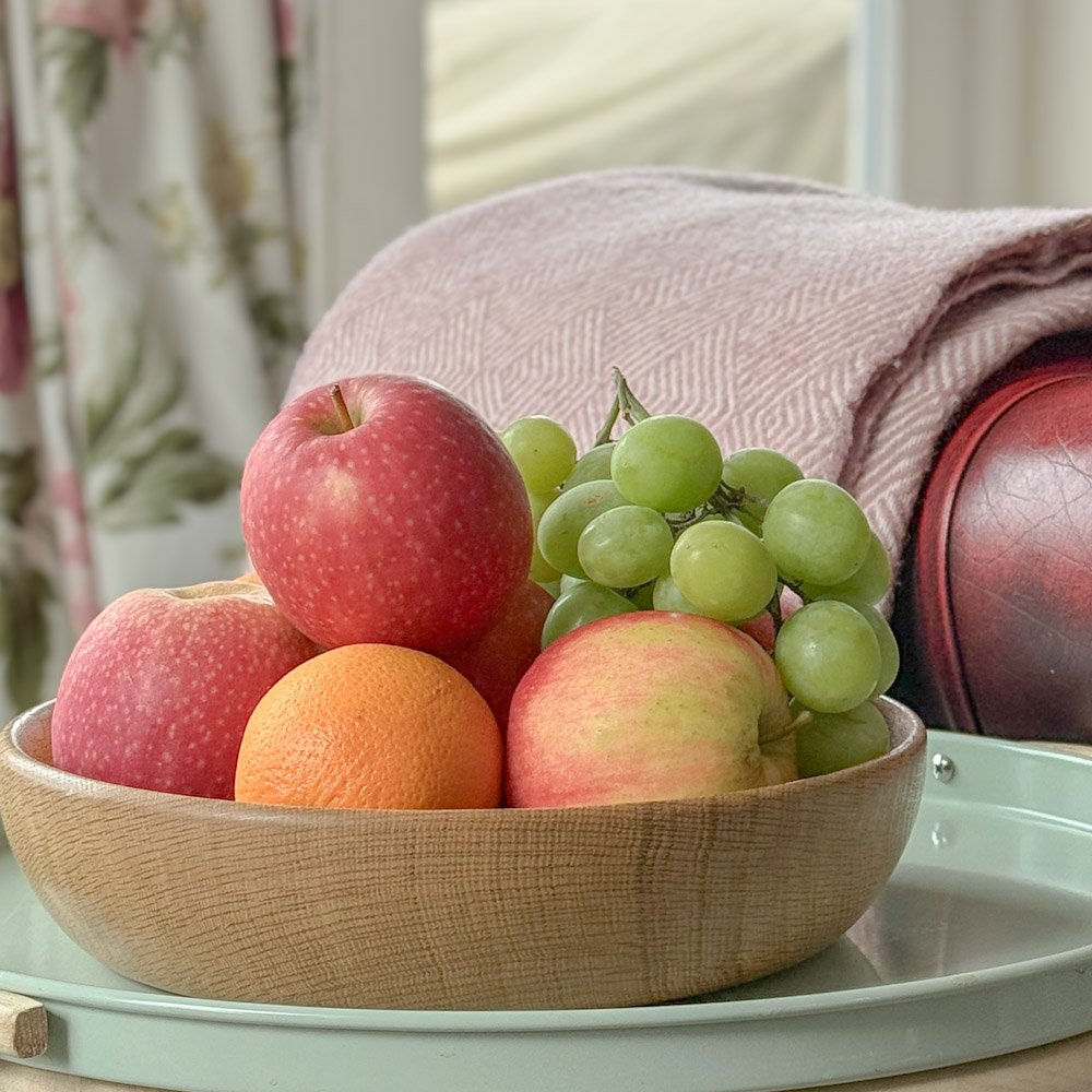 natural oak serving board filled with fresh fruit, apples, oranges and grapes, the bowl is on a small metal side table next to a leather chesterfield chair, a pink wool blanket is laid neatly over the arm of the chair, laura ashley curtains hang at the french doors behind. the oak bowl is handmade in Scotland