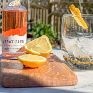 Scottish Gin Whisky Cask G&T Board on an outside table in the sunshine. A bottle of Great glen Gin is behind the board and a sliced lemon is on it. A glass with ice and lemon to the right. You can see the garden fence and some shrubs behind. the G& T board is Scottish Made