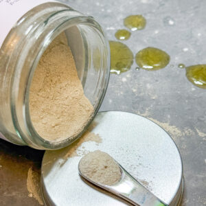 Ola Ròs Skincare Refine Purifying Facial Masque glass jar with pink label Ola Ròs Skincare Refine Purifying facial Masque the jar is laid on is side on the metal lid and some of the powder has spilled out. A small metal cosmetic scoop is sat on the lid with a little powder on it a few drops of balance oil cleanser are on the grey worktop above it
