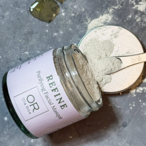 Ola Ròs Skincare Refine Purifying Facial Masque glass jar with pink label Ola Ròs Skincare Refine Purifying facial Masque the jar is laid on is side on the metal lid and some of the powder has spilled out. A small metal cosmetic scoop is sat on the lid with a little powder on it a few drops of balance oil cleanser are on the grey worktop above it