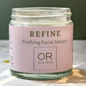 Ola Ròs Skincare Refine Purifying Facial Masque glass jar with pink label Ola Ròs Skincare Refine Purifying Facial Masque some of the powdered masque is spilled on the grey worktop it is stood on. A green background behind. handmade in Scotland