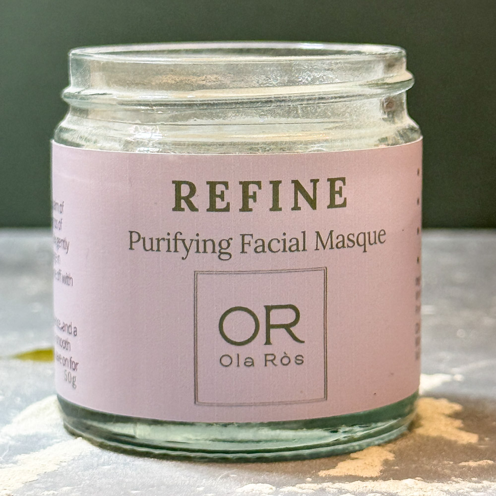 Ola Ròs Skincare Refine Purifying Facial Masque glass jar with pink label Ola Ròs Skincare Refine Purifying Facial Masque some of the powdered masque is spilled on the grey worktop it is stood on. A green background behind. handmade in Scotland