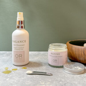Ola Ròs Skincare Refine Purifying Facial Masque glass jar with pink label Ola Ròs Skincare Refine Purifying facial Masque and a glass whie bottle of Ola Ròs Skincare Balance Oil Cleanser a few drops of balance are on the worktop and the lid of the refine powder is off. A small cosmetic scoop with some powder on it behind all the products is a small wooden bowl the background is green