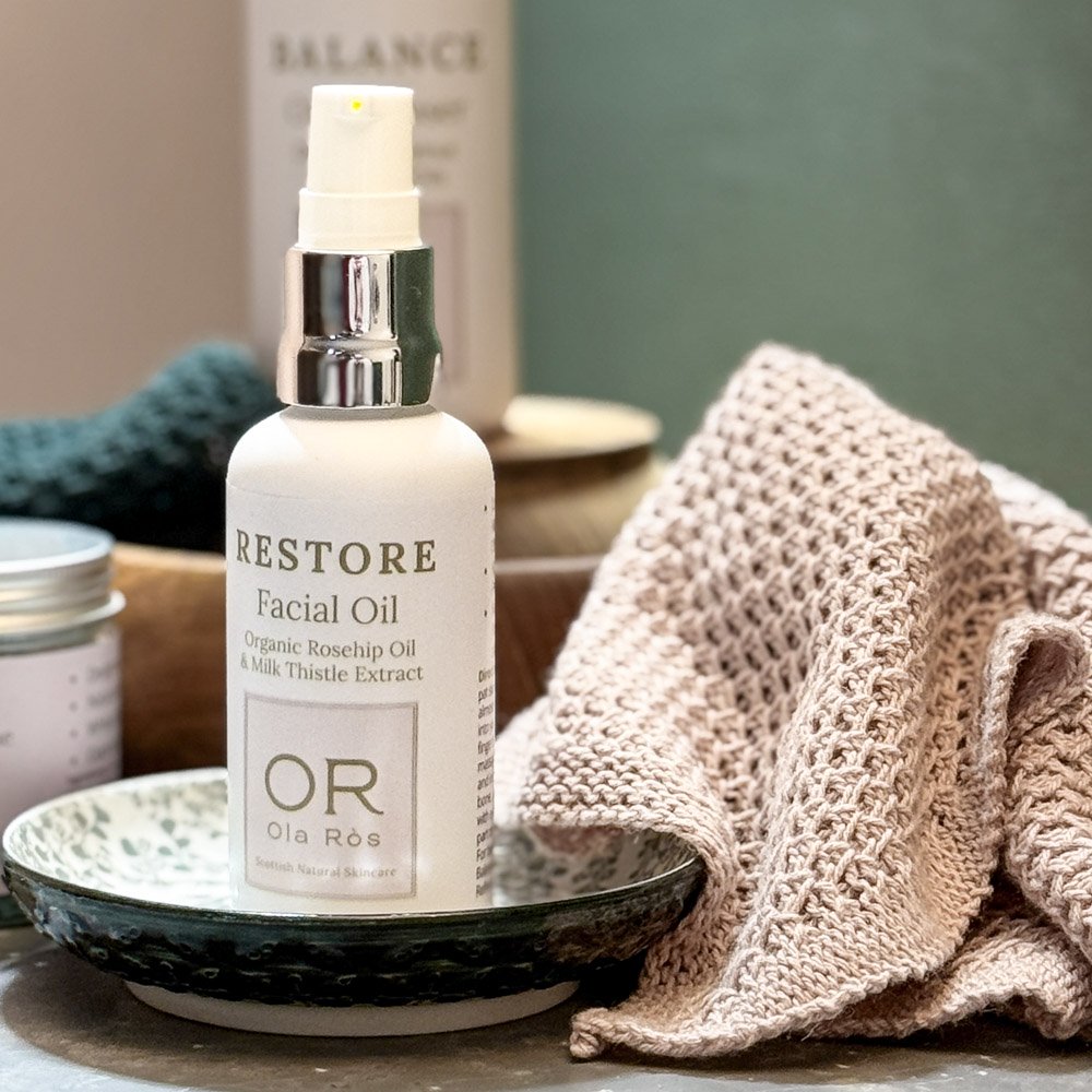 Restore Facial Oil by Ola Ròs skincare in a white matt glass bottle stood on a grey tile with a green and pink background and a pink linen face cloth