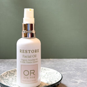 Restore Facial Oil by Ola Ròs skincare in a white matt glass bottle stood on a grey tile with a green background. Handmade in Scotland