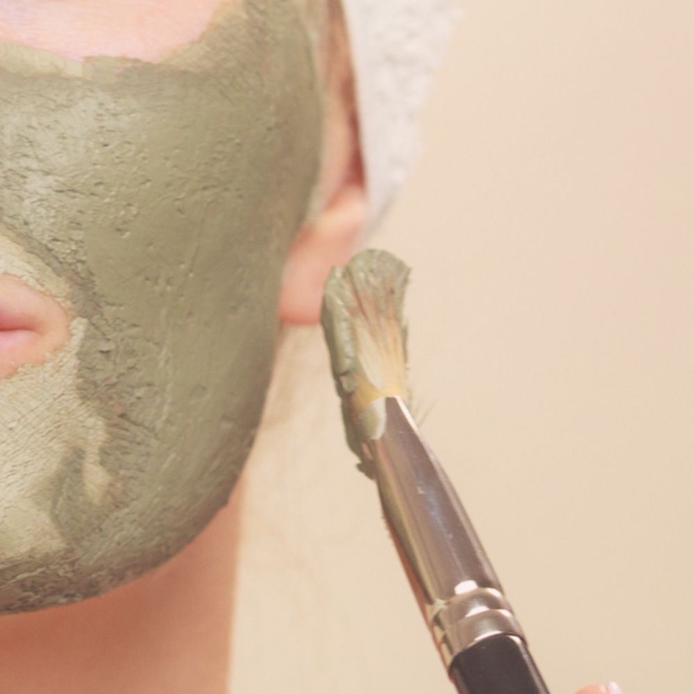 lady applying refine facial masque with a makeup brush to her face only the cheek and a little bit of ear and tiny bit of the mouth can be seen in the image and the make up brush is held slightly away from her face.