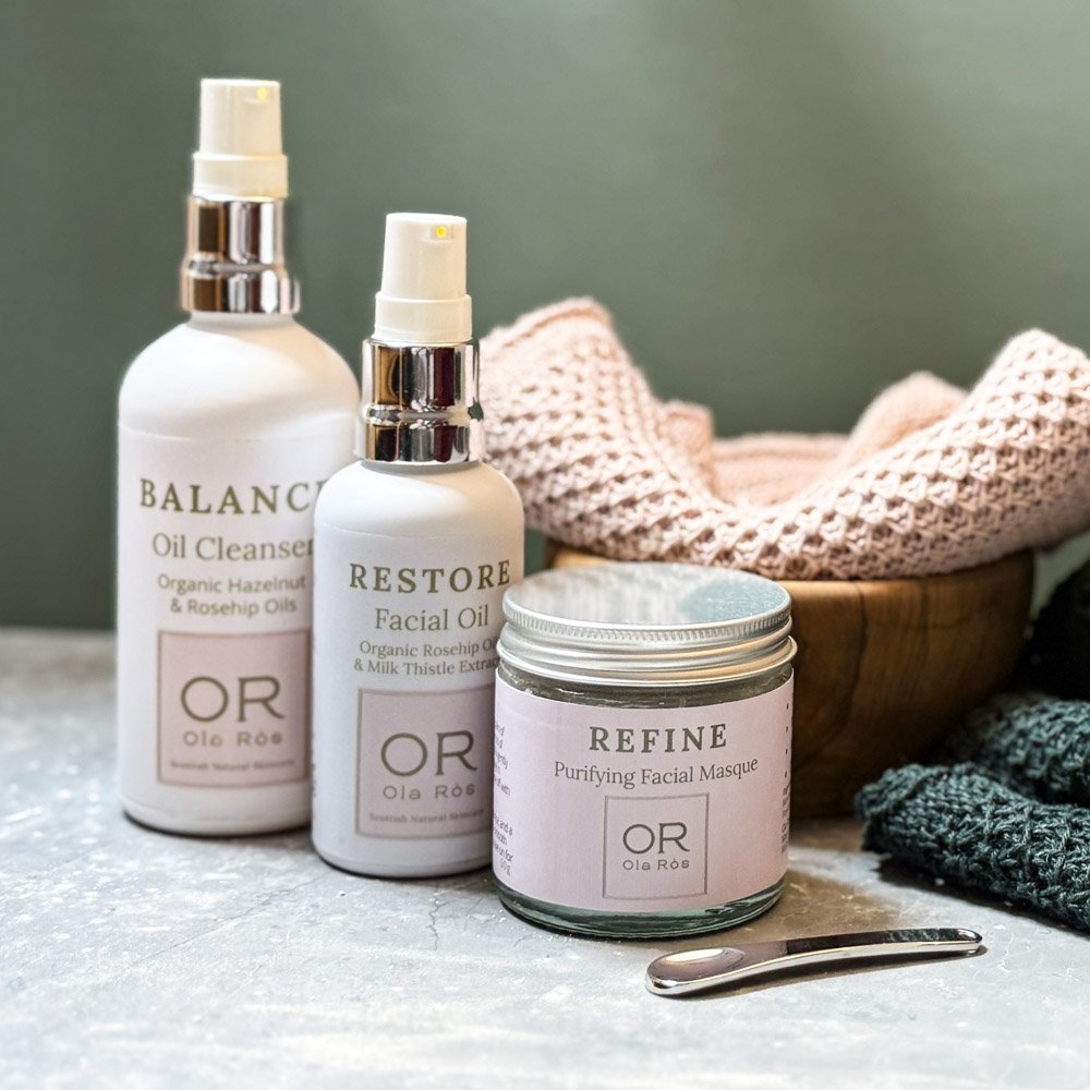 the complete ritual with linen face cloth set out on a grey worktop a white bottle of balance oil cleanser, a white bottle of restore facial oil and a jar of refine purifying facial masque are nest to a wooden bowl that has a pink linen facecloth inside it. a small cosmetic scoop sits beside the bottles