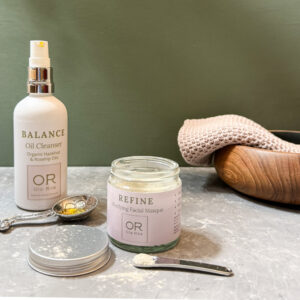 Ola Ròs Skincare Refine Purifying Facial Masque glass jar with pink label Ola Ròs Skincare Refine Purifying facial Masque and a glass whie bottle of Ola Ròs Skincare Balance Oil Cleanser a few drops of balance are on the worktop and the lid of the refine powder is off. A small cosmetic scoop with some powder on it behind all the products is a small wooden bowl the background is green