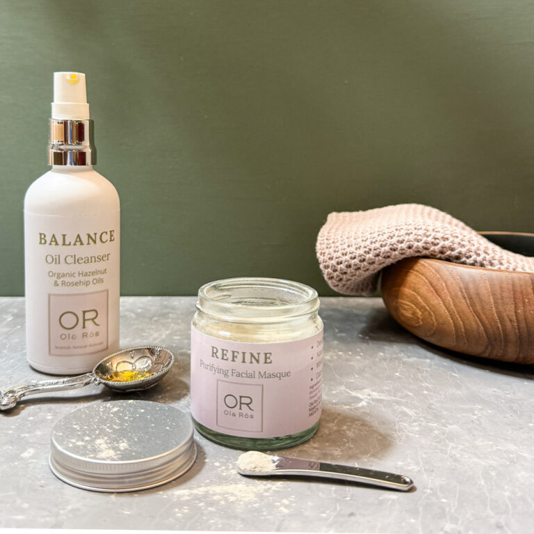 Ola Ròs Skincare Refine Purifying Facial Masque glass jar with pink label Ola Ròs Skincare Refine Purifying facial Masque and a glass whie bottle of Ola Ròs Skincare Balance Oil Cleanser a few drops of balance are on the worktop and the lid of the refine powder is off. A small cosmetic scoop with some powder on it behind all the products is a small wooden bowl the background is green