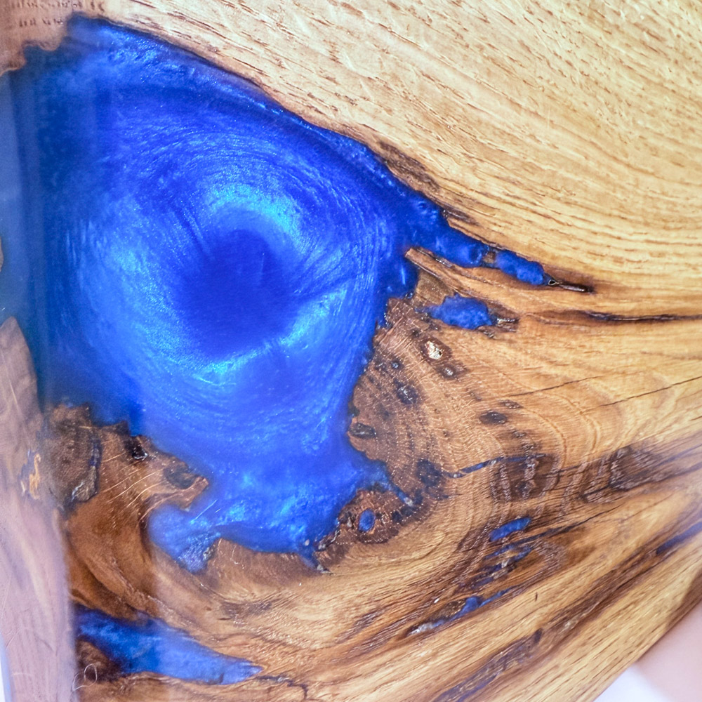 Oak river board standing upright on a pink tabletop with a green tiled background, a close up showing the electric-blue resin swirl on the left side and the rich oak grain,