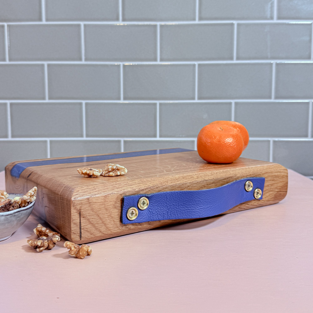 Handcrafted oak river board with an electric-blue inlay and blue leather handle, resting on a pink tabletop with a green tiled backdrop, styled with an orange and a few walnuts