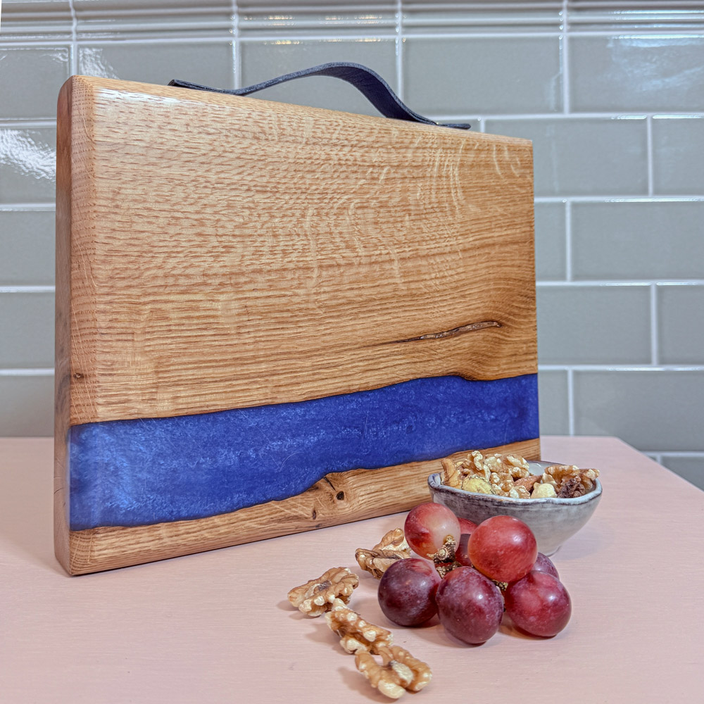 Oak river board standing upright on a pink tabletop with a green tiled background, electric-blue resin stripe across the length of the board, styled with a bowl of walnuts and a small bunch of grapes