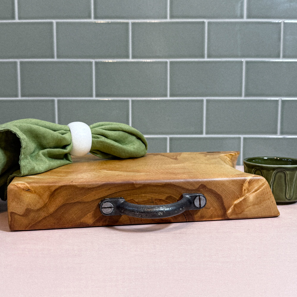 Birch Wood Board with reclaimed metal handle laid on its side with a green napkin and ring laid on top. the background it fired earth green tiles and the tabletop is pink the birch board was handmade in scotland from local Highland wood