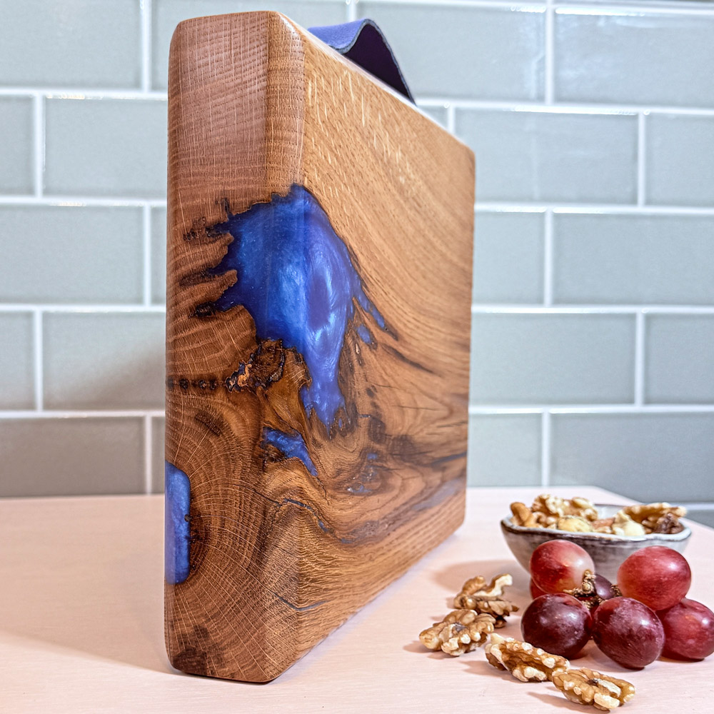 Oak river board standing upright on a pink tabletop with a green tiled background, showing the electric-blue resin swirl on the left side and the rich oak grain, styled with a bowl of walnuts and a small bunch of grapes