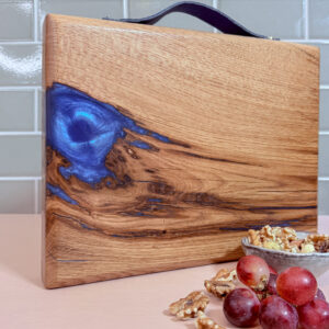 Handmade in Scotland and Scottish Made Oak river board standing upright on a pink tabletop with a green tiled background, showing the electric-blue resin swirl on the left side and the rich oak grain, styled with a bowl of walnuts and a small bunch of grapes