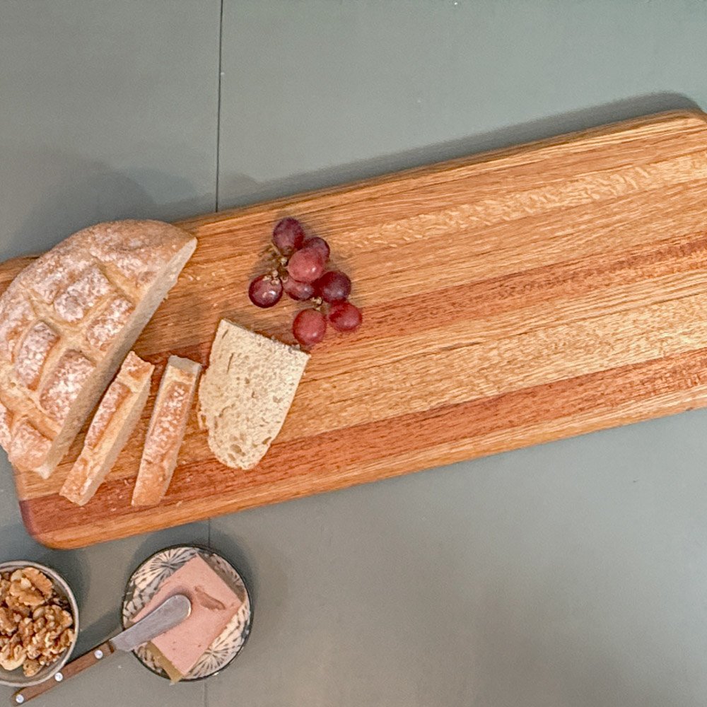 subtly striped handmade oak and mahogany chopping board made from oak and mahogany laying on a green table styled with bread grapes and pate with a green tiled background