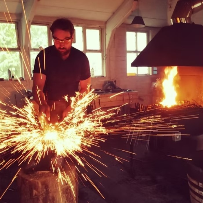 Derro Gilmour forging at highland Forge. Sparks are flying out of the metal as he hammers the hot piece straight from the forge. you can see the chimney hood of the coal forge behind him.