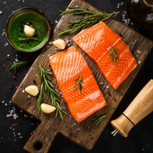 Two pieces of salmon laid out on a board prior to cooking, seasoning such as salt and pepper and rosemary and garlic surround it. salmon is full of omega-3 fatty acids that bring health benefits to our skin cells