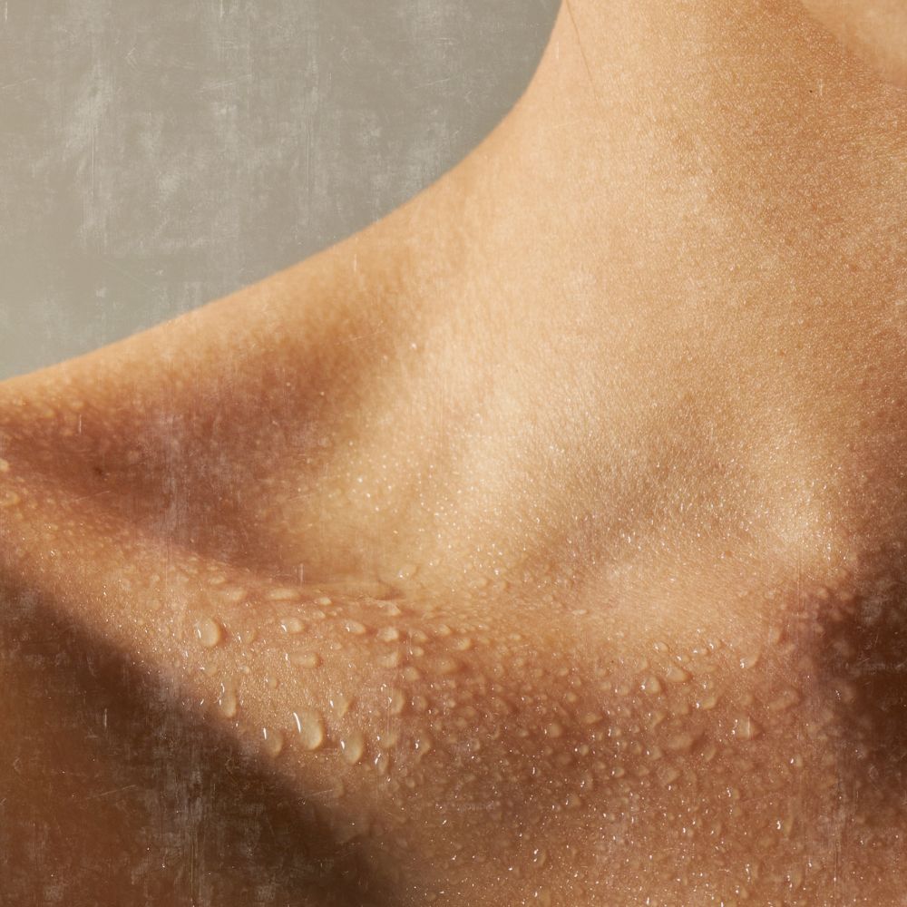 image of a female collarbone and neck and partial shoulder that is covered in soft shadow and water droplets to depict the idea of well hydrated skin