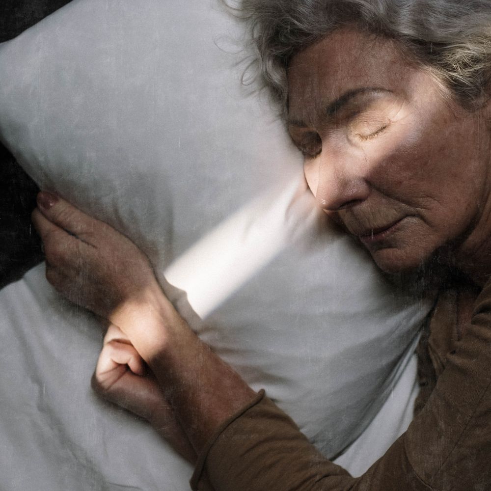 older lady laying in bed asleep her bedding is white, her eyes are close and her hands are crossed, a shaft of soft evening light comes across her face