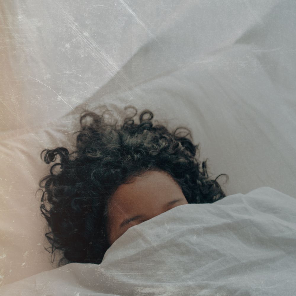 Lady with dark hair sleeping in bed then sheets and pillows are white she is tucked under the quilt so that only her eyebrows and forehead are above the covers