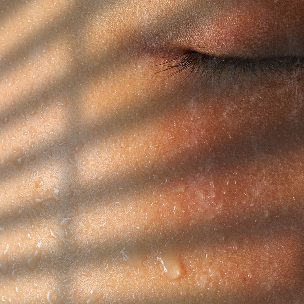 image of a lady's face she has her eyes closed, water droplets are on her skin and a shadow is being cast by a blind
