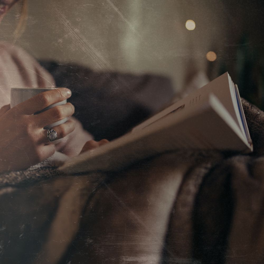 lady sat irelaxing in the evening reading a book, she has a cup of tea in her hand and a blanket over her knees we cant see her face in the image, its really a close up of the book, hands and cup depicting relaxing evening routines for good sleep hygiene