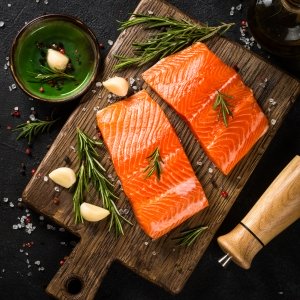 Two pieces of salmon laid out on a board prior to cooking, seasoning such as salt and pepper and rosemary and garlic surround it. salmon is full of omega-3 fatty acids that bring health benefits to our skin cells