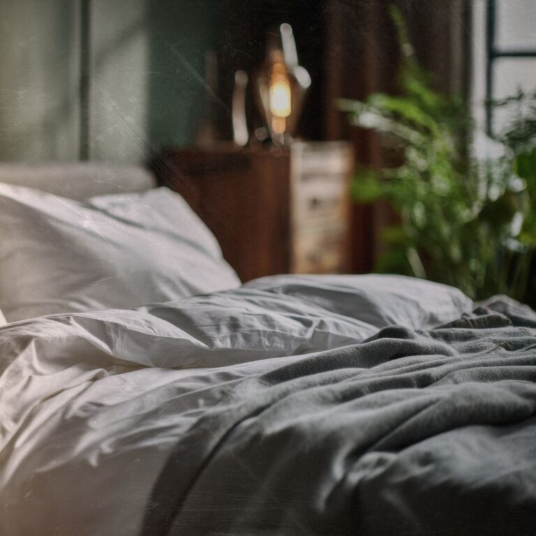 crumbled linen sheets on a bed in the soft light of the evening, a chest of drawers is beside the bed with a lamp on top and a houseplant beside this image is being sued to show the importance of a good sleep cycle in our skin health