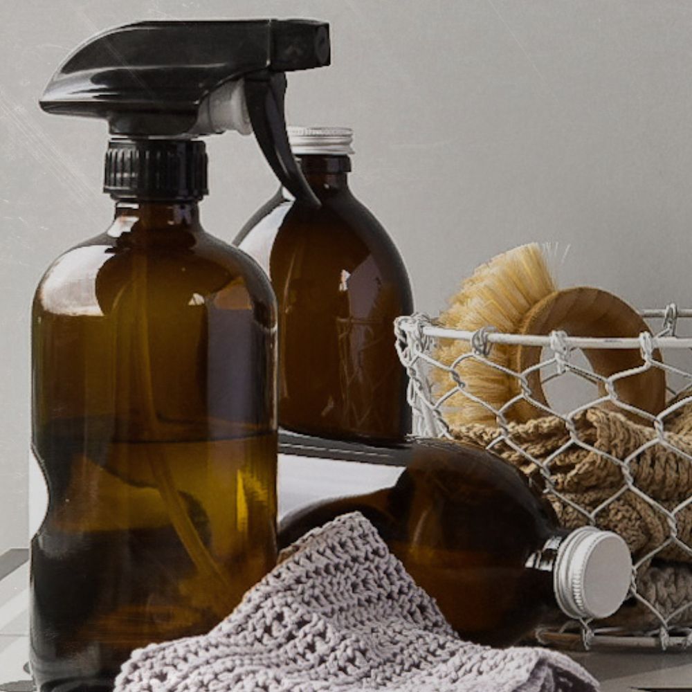All-Purpose household spray in a glass amber bottle with a trigger spray . a basket of dishcloths is offset in the image and two more amber glass bottles are also in the image, a hand crocheted dishcloth is at the front of the image.