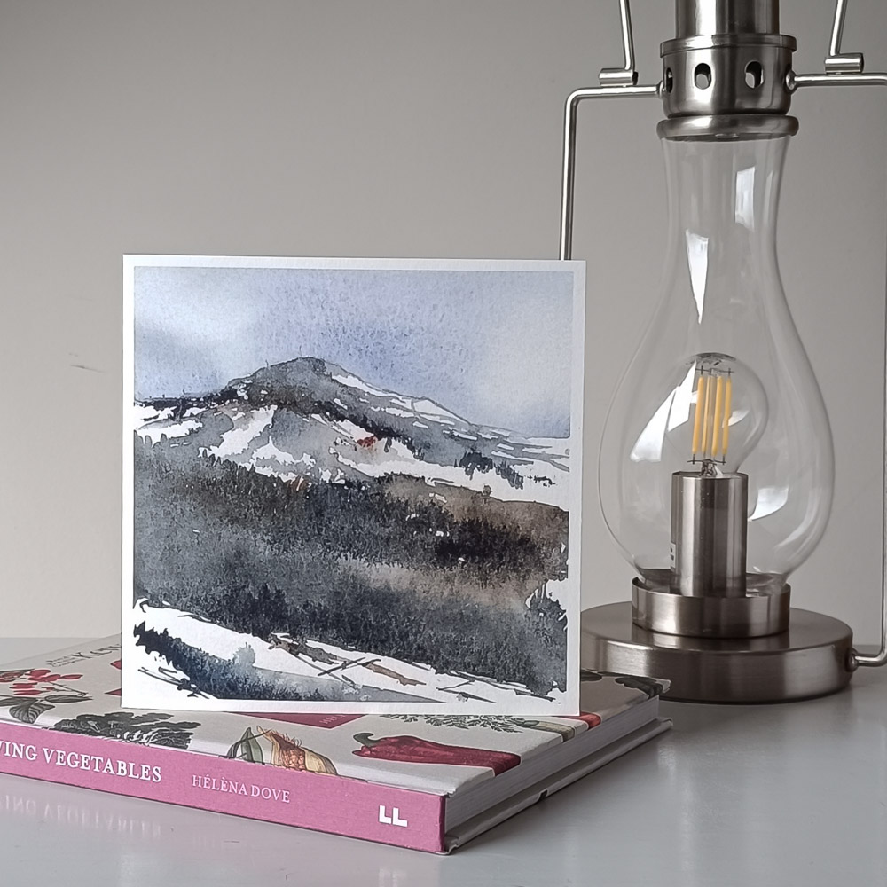 a staged image of the square greeting card a watercolour painting by julia anne of highland landscape Suidh Ghuirmain stood on a kew gardens book and a lantern lamp