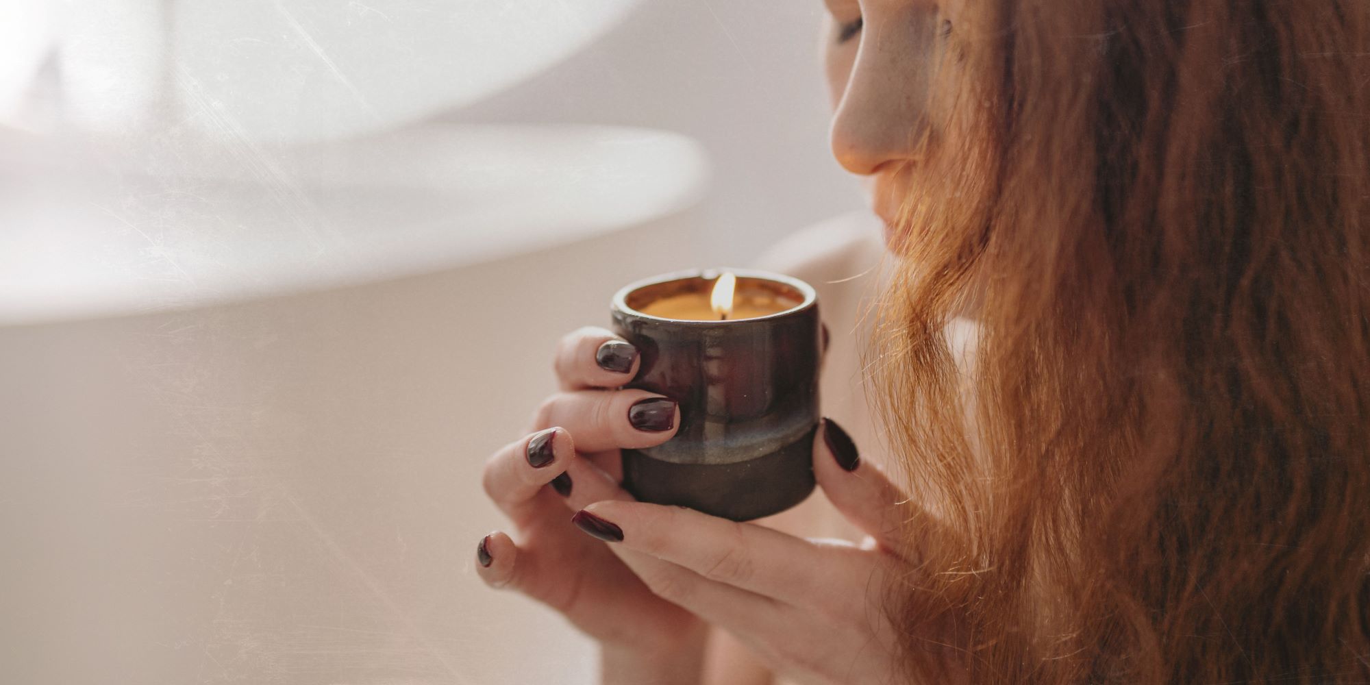 a red headed lady sniffing a burning container candle in a light background bathroom